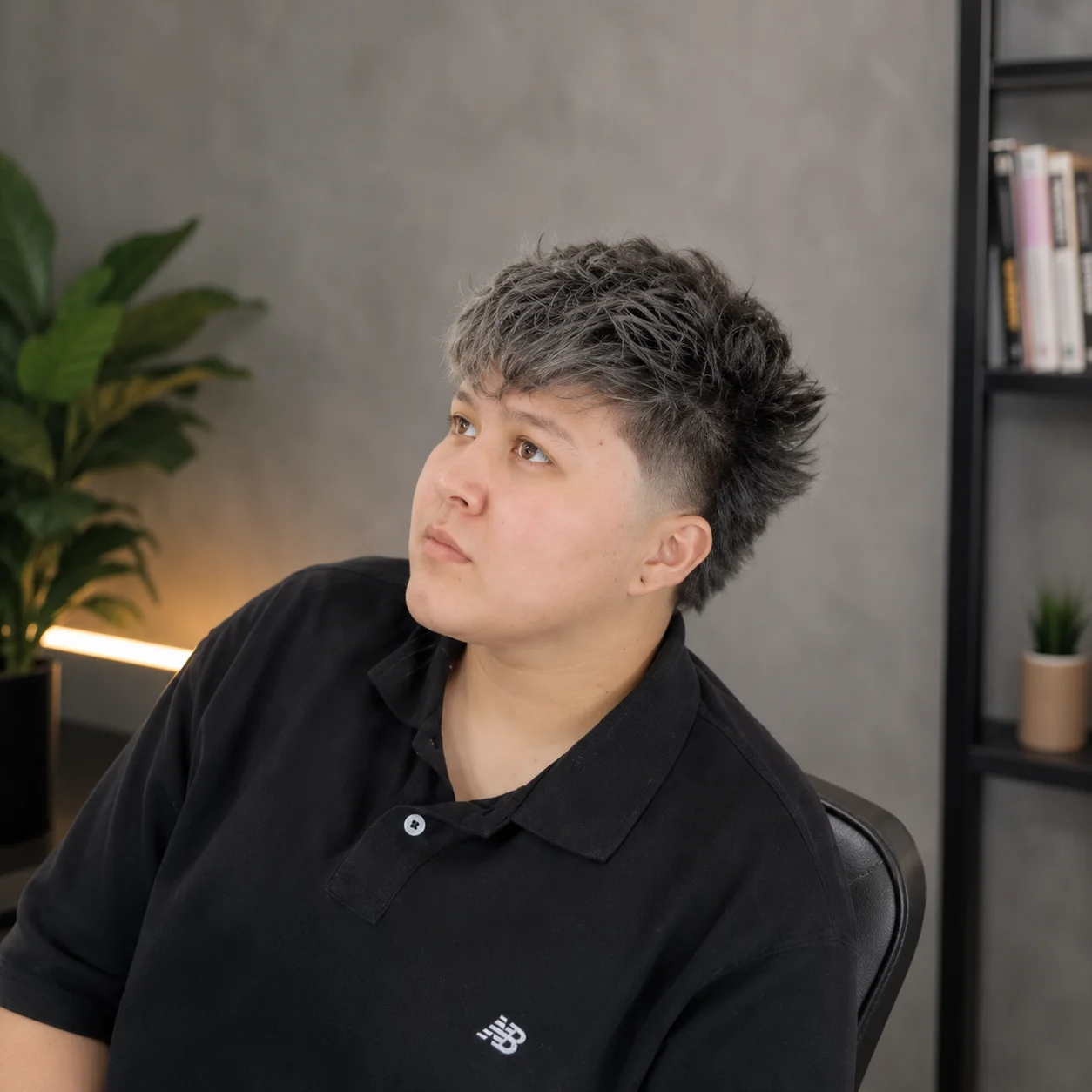 Textured short haircut with faded sides on a young person wearing a black polo shirt, looking slightly upward in a modern indoor studio setting with a soft blurred background.
