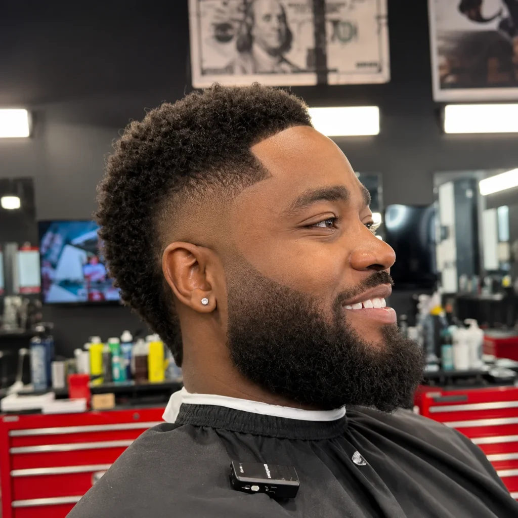 Side profile of a man with curly textured top and low taper fade haircut, sharp lineup with a full beard, styled in a professional barbershop setting.